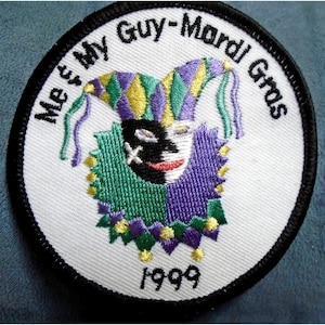 May include: Embroidered patch with the text "Meet My Guy-Mardi Gras" and the year 1999. The patch features a jester's face with a black mask, a purple and yellow hat, and a green collar. The patch has a white background and a black border.
