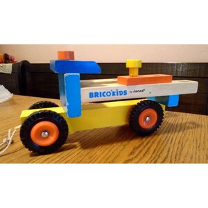 May include: A wooden toy truck with a blue cab, yellow bed, and orange wheels. The truck has a wooden platform with the words "BRICO'KIDS by Janod" printed on it. The truck is a construction toy for children.