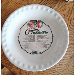 May include: A white ceramic pie dish with a scalloped rim. The center features a printed recipe for Country Apple Pie, including ingredients and instructions, framed by a brown branch illustration with two red apples and green leaves at the top.