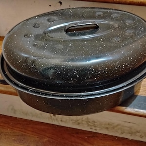 May include: Black oval roasting pan with a speckled enamel finish. The pan has a lid with a handle and is sitting on a wooden surface. The pan has two handles on the sides.