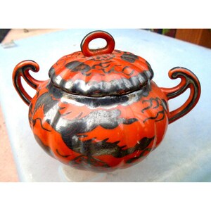 May include: A vintage, round sugar bowl with a lid. The bowl is a vibrant orange with black and silver accents. It has two curved handles and a loop handle on the lid. The design appears to be hand-painted.