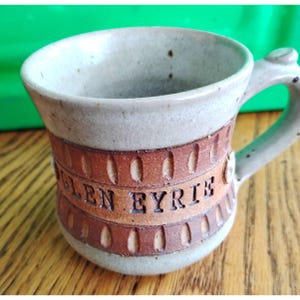 May include: A handmade ceramic mug with a light beige exterior and a brown band featuring the text "GLEN EYRIE." The mug has a handle and is suitable for hot or cold beverages. The mug is approximately 3.5 inches tall.
