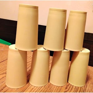 May include: A stack of eight beige, cylindrical cups. The cups are arranged in a pyramid shape, with three cups on the bottom row, two in the middle, and one on top. The cups are made of a light-colored material and have a simple, minimalist design.