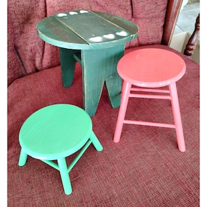 May include: Three small, painted wooden stools in shades of green and coral. The largest stool is dark green with a round top and three legs. The smallest stool is light green, and the middle stool is coral pink.
