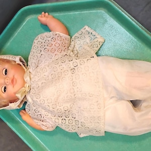 May include: A doll with blonde hair and painted facial features wearing a white lace bonnet, a white lace short-sleeved top, white trousers, and matching lace booties. The doll is positioned in a supine orientation with arms extended outward.