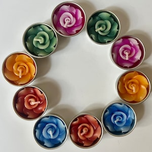 May include: Ten tealight candles in metal containers, each with a different colored wax rose design. The candles are arranged in a circle on a white surface.