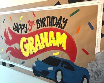 May include: A rectangular brown paper banner featuring the text Happy 3rd Birthday Graham in black and yellow lettering. A red flame graphic sits behind the name, and a blue sports car with grey smoke is illustrated at the bottom.