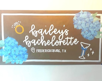 May include: A rectangular brown sign with a white border featuring the hand-painted text "bailey's bachelorette" and "FREDERICKSBURG, TX". The design includes a gold ring, a martini glass, and clusters of blue hydrangea flowers.