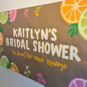 May include: A brown sign featuring the white text KAITLYN'S BRIDAL SHOWER and the yellow script text she found her main squeeze. The design is decorated with painted illustrations of orange, lime, and pink grapefruit slices and small flowers.