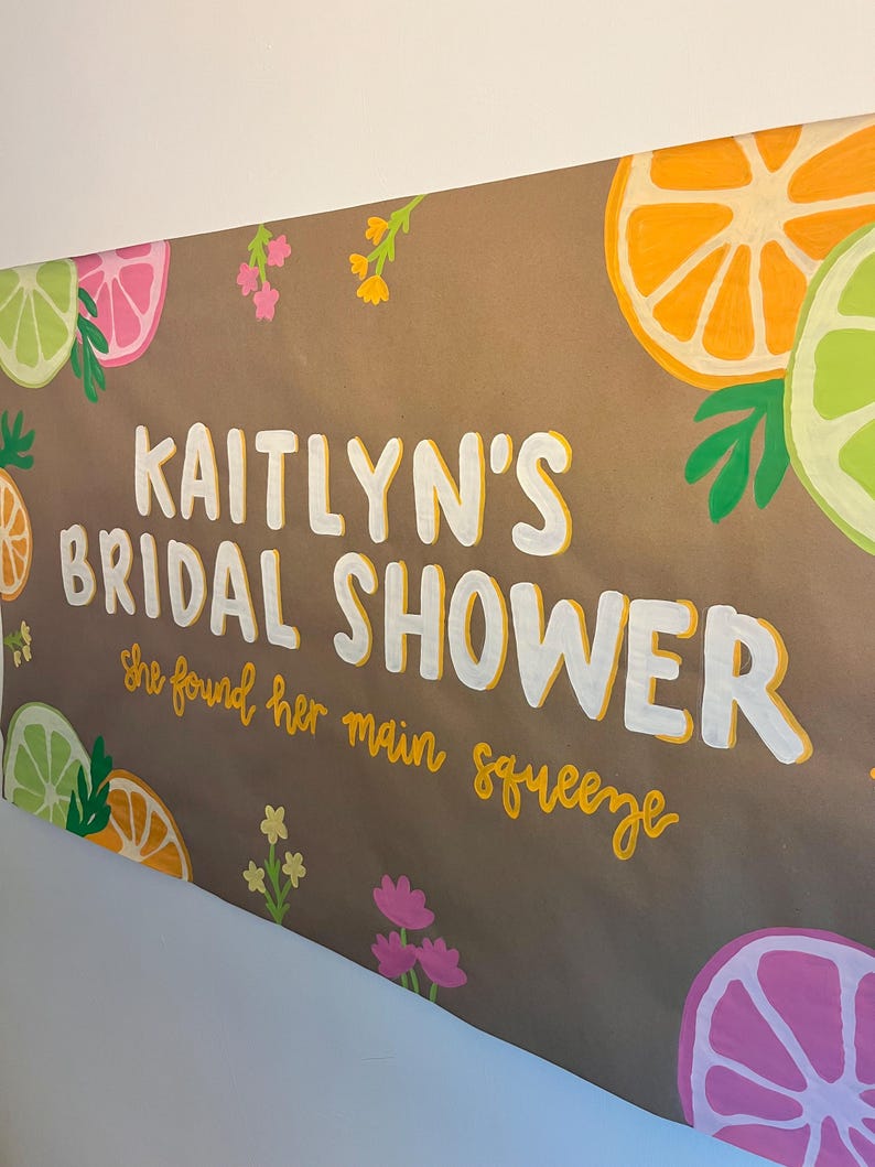 May include: A brown sign featuring the white text KAITLYN'S BRIDAL SHOWER and the yellow script text she found her main squeeze. The design is decorated with painted illustrations of orange, lime, and pink grapefruit slices and small flowers.