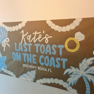 May include: A brown paper banner featuring the hand-painted text Kate's LAST TOAST ON THE COAST ROSEMARY BEACH, FL in white and light blue. The design includes white pearl strands, light blue seashells, a gold ring, and a palm tree.