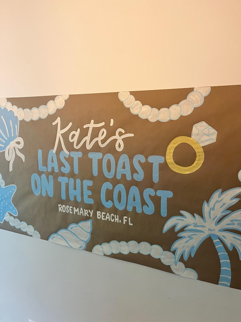 May include: A brown paper banner featuring the hand-painted text Kate's LAST TOAST ON THE COAST ROSEMARY BEACH, FL in white and light blue. The design includes white pearl strands, light blue seashells, a gold ring, and a palm tree.