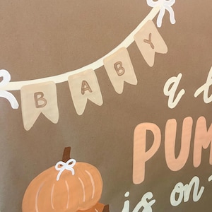 May include: A brown sign featuring a painted banner with the word BABY in white and brown letters. Below the banner, two orange pumpkins are painted with white bows on their stems. The text PUMPKIN is partially visible in large orange letters.