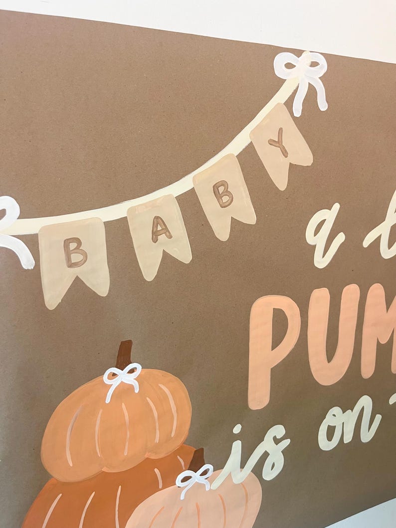 May include: A brown sign featuring a painted banner with the word BABY in white and brown letters. Below the banner, two orange pumpkins are painted with white bows on their stems. The text PUMPKIN is partially visible in large orange letters.