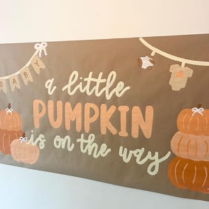 May include: A rectangular brown banner featuring the text "a little PUMPKIN is on the way" in white and orange. The design includes illustrations of orange pumpkins, a string of baby clothes, and a pennant banner spelling "BABY" in white.