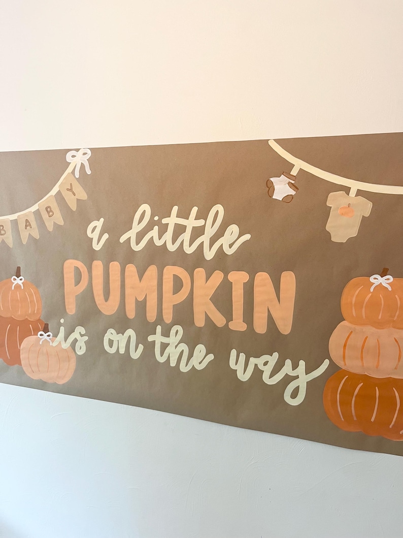 May include: A rectangular brown banner featuring the text "a little PUMPKIN is on the way" in white and orange. The design includes illustrations of orange pumpkins, a string of baby clothes, and a pennant banner spelling "BABY" in white.