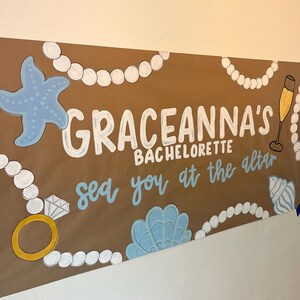 May include: A brown paper banner featuring the white text GRACEANNA'S BACHELORETTE and the blue text sea you at the altar. The design includes illustrations of white pearl necklaces, a blue starfish, a gold ring with a diamond, a champagne flute, and blue seashells.
