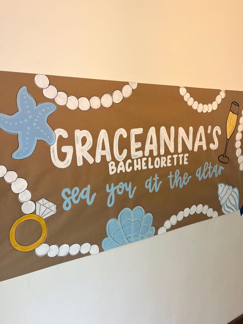 May include: A brown paper banner featuring the white text GRACEANNA'S BACHELORETTE and the blue text sea you at the altar. The design includes illustrations of white pearl necklaces, a blue starfish, a gold ring with a diamond, a champagne flute, and blue seashells.