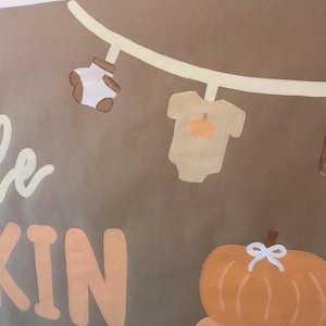May include: A brown sign featuring a painted clothesline with a pair of white baby socks and a beige baby bodysuit with a pumpkin graphic. Below the clothesline, the text KIN is visible in large peach letters, alongside a stack of two orange pumpkins.