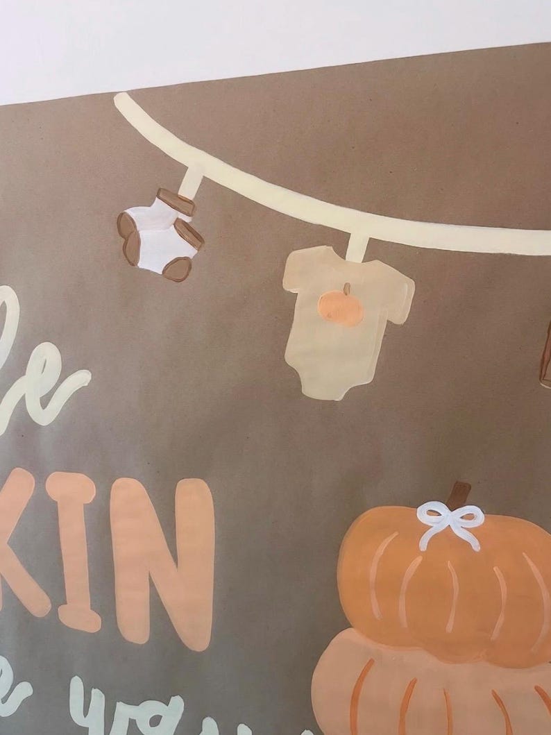 May include: A brown sign featuring a painted clothesline with a pair of white baby socks and a beige baby bodysuit with a pumpkin graphic. Below the clothesline, the text KIN is visible in large peach letters, alongside a stack of two orange pumpkins.