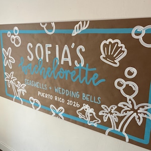 May include: A rectangular brown paper banner featuring a light blue border and white hand-painted illustrations of seashells, palm trees, bubbles, and wedding bells. The text reads: SOFIA'S bachelorette SEASHELLS + WEDDING BELLS PUERTO RICO 2026.