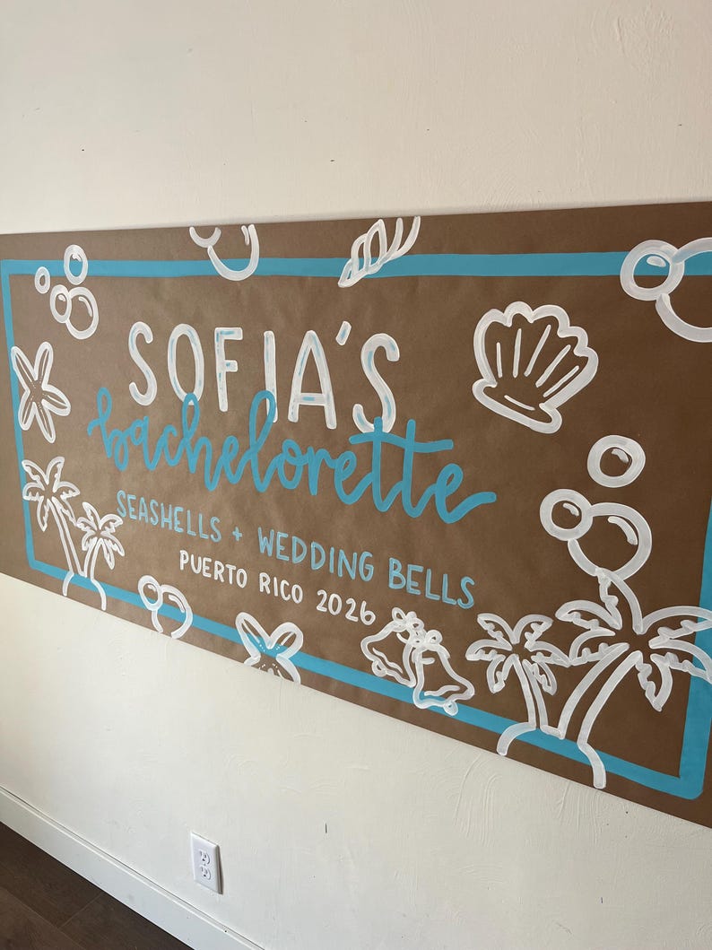 May include: A rectangular brown paper banner featuring a light blue border and white hand-painted illustrations of seashells, palm trees, bubbles, and wedding bells. The text reads: SOFIA'S bachelorette SEASHELLS + WEDDING BELLS PUERTO RICO 2026.
