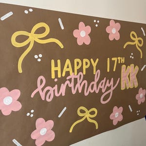 May include: A brown rectangular poster featuring the hand-painted text HAPPY 17TH birthday KK in yellow and pink. The design is decorated with yellow bows, pink flowers with white centers, and small white dash and dot accents scattered throughout.