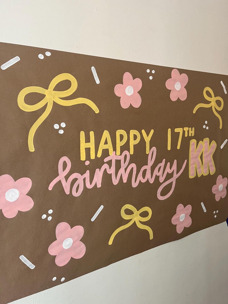 May include: A brown rectangular poster featuring the hand-painted text HAPPY 17TH birthday KK in yellow and pink. The design is decorated with yellow bows, pink flowers with white centers, and small white dash and dot accents scattered throughout.