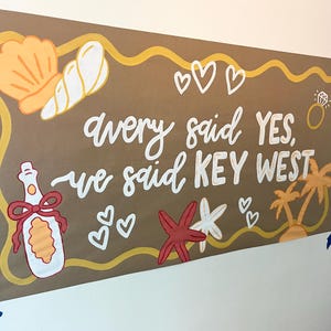 May include: A rectangular brown sign featuring the white hand-painted text Avery said YES, we said KEY WEST. The design includes illustrations of a seashell, a champagne bottle with a red bow, a diamond ring, hearts, a starfish, and palm trees.