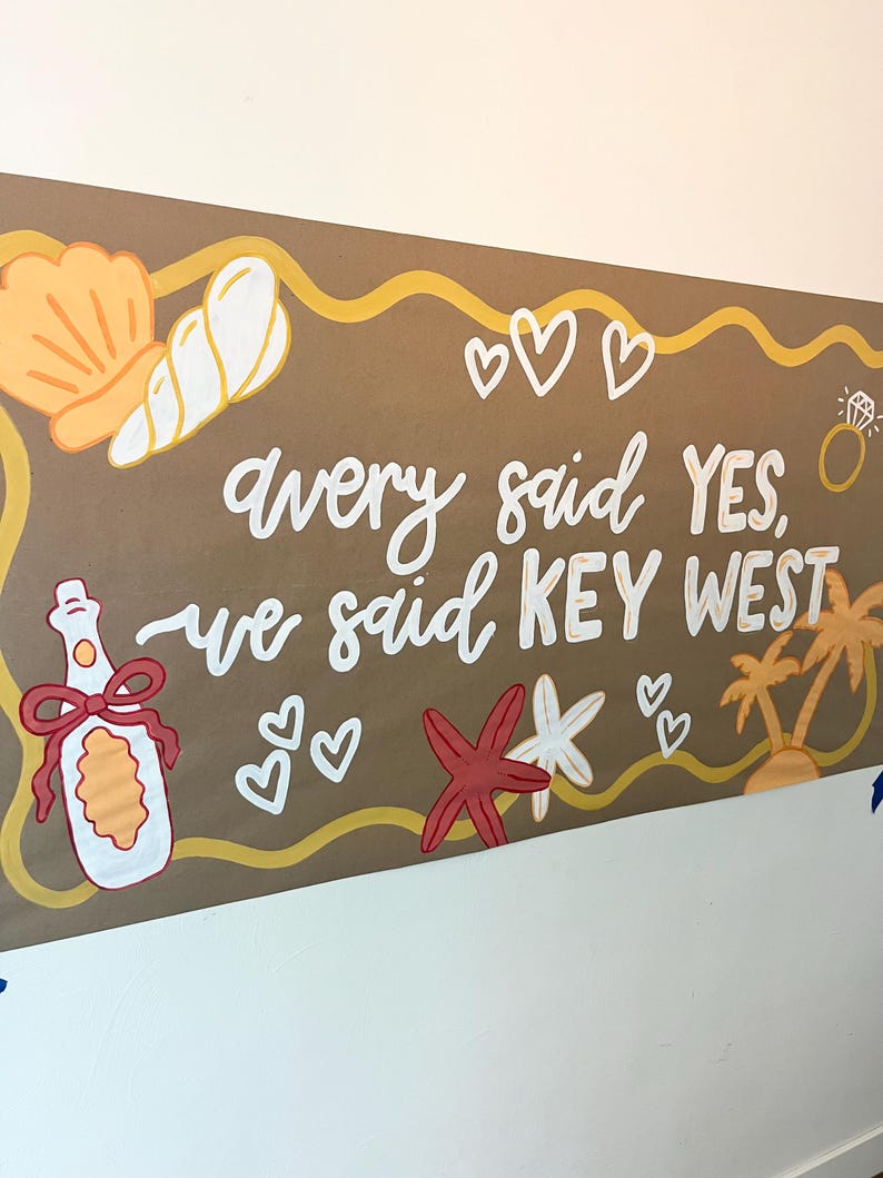 May include: A rectangular brown sign featuring the white hand-painted text Avery said YES, we said KEY WEST. The design includes illustrations of a seashell, a champagne bottle with a red bow, a diamond ring, hearts, a starfish, and palm trees.