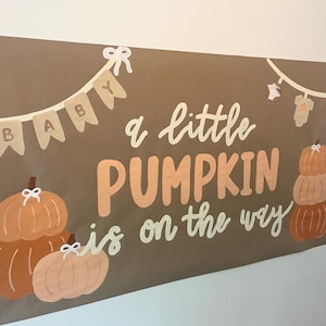 May include: A brown rectangular banner featuring the text "a little PUMPKIN is on the way" in white and orange. The design includes illustrations of orange pumpkins, a bunting with the word "BABY", and a clothesline with baby onesies.
