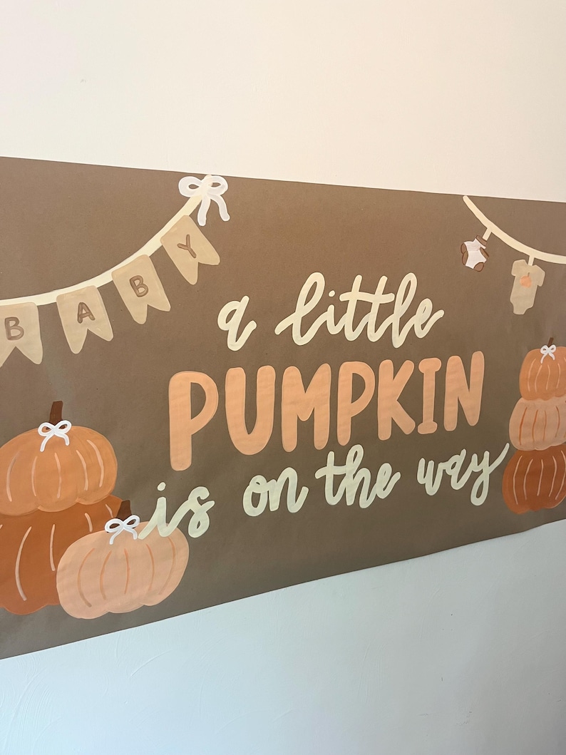 May include: A brown rectangular banner featuring the text "a little PUMPKIN is on the way" in white and orange. The design includes illustrations of orange pumpkins, a bunting with the word "BABY", and a clothesline with baby onesies.