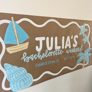 May include: A brown paper banner featuring white wavy borders and hand-painted illustrations including a blue sailboat, a seashell, two starfish, and a palm tree. The text reads JULIA'S bachelorette weekend, CHARLESTON, SC - OCT 2025 in white and blue.