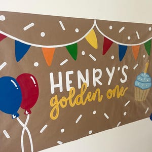 May include: A brown paper banner featuring the white text "HENRY'S" and the yellow script text "golden one". The design includes colorful triangular bunting, white confetti dots, a blue and red balloon pair, and a blue cupcake with a candle.