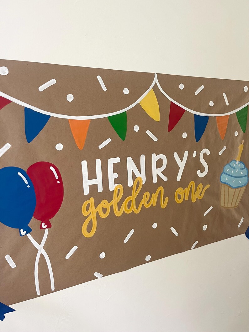 May include: A brown paper banner featuring the white text "HENRY'S" and the yellow script text "golden one". The design includes colorful triangular bunting, white confetti dots, a blue and red balloon pair, and a blue cupcake with a candle.