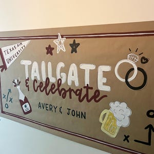 May include: A large brown paper banner featuring the hand-painted text Tailgate & Celebrate and Avery & John. Illustrations include a Texas A&M University pennant, stars, two interlocking rings, a champagne bottle, and a mug of beer with foam.