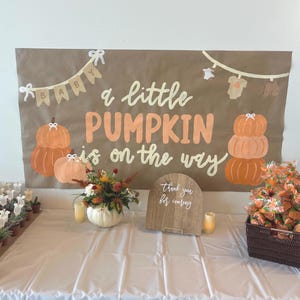May include: A brown rectangular banner with the text "a little PUMPKIN is on the way" in white and orange lettering. The banner features illustrations of pumpkins, baby clothes on a clothesline, and a "BABY" bunting. It is displayed above a table.