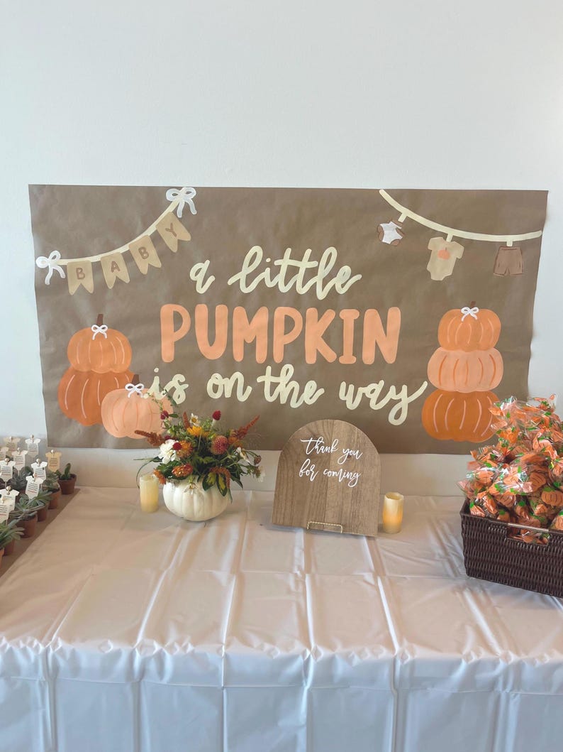 May include: A brown rectangular banner with the text "a little PUMPKIN is on the way" in white and orange lettering. The banner features illustrations of pumpkins, baby clothes on a clothesline, and a "BABY" bunting. It is displayed above a table.