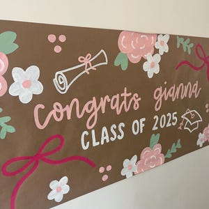 May include: A brown paper banner featuring hand-painted floral designs in pink, white, and green, along with a diploma and graduation cap illustration. The text reads "congrats gianna" in pink script and "CLASS OF 2025" in white block letters.