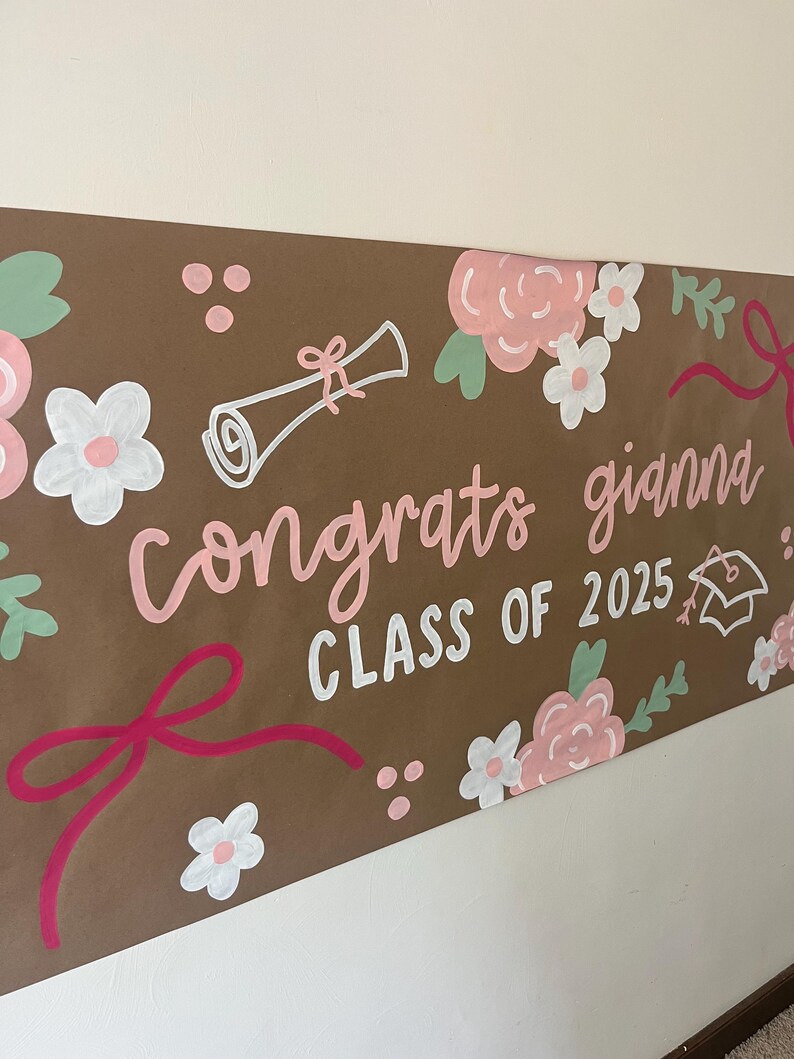 May include: A brown paper banner featuring hand-painted floral designs in pink, white, and green, along with a diploma and graduation cap illustration. The text reads "congrats gianna" in pink script and "CLASS OF 2025" in white block letters.