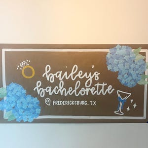 May include: A rectangular brown sign with a white border featuring the hand-painted white text "bailey's bachelorette" and "FREDERICKSBURG, TX". The design includes blue hydrangea clusters, a gold ring icon, and a white martini glass graphic.