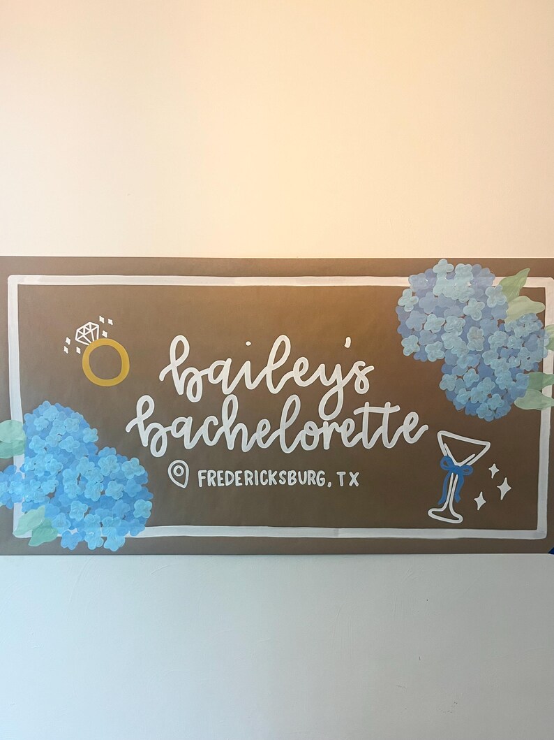 May include: A rectangular brown sign with a white border featuring the hand-painted white text "bailey's bachelorette" and "FREDERICKSBURG, TX". The design includes blue hydrangea clusters, a gold ring icon, and a white martini glass graphic.