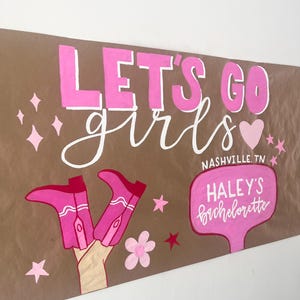 May include: A brown paper sign featuring the pink text LET'S GO girls, a pink heart, and two pink cowboy boots. It includes a pink speech bubble with the text NASHVILLE, TN HALEY'S Bachelorette, decorated with pink stars and a flower.