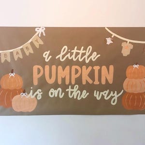 May include: A rectangular brown banner featuring the text "a little PUMPKIN is on the way" in white and orange. The design includes illustrations of orange pumpkins with white bows and a string of baby clothes and pennants with the word "BABY".