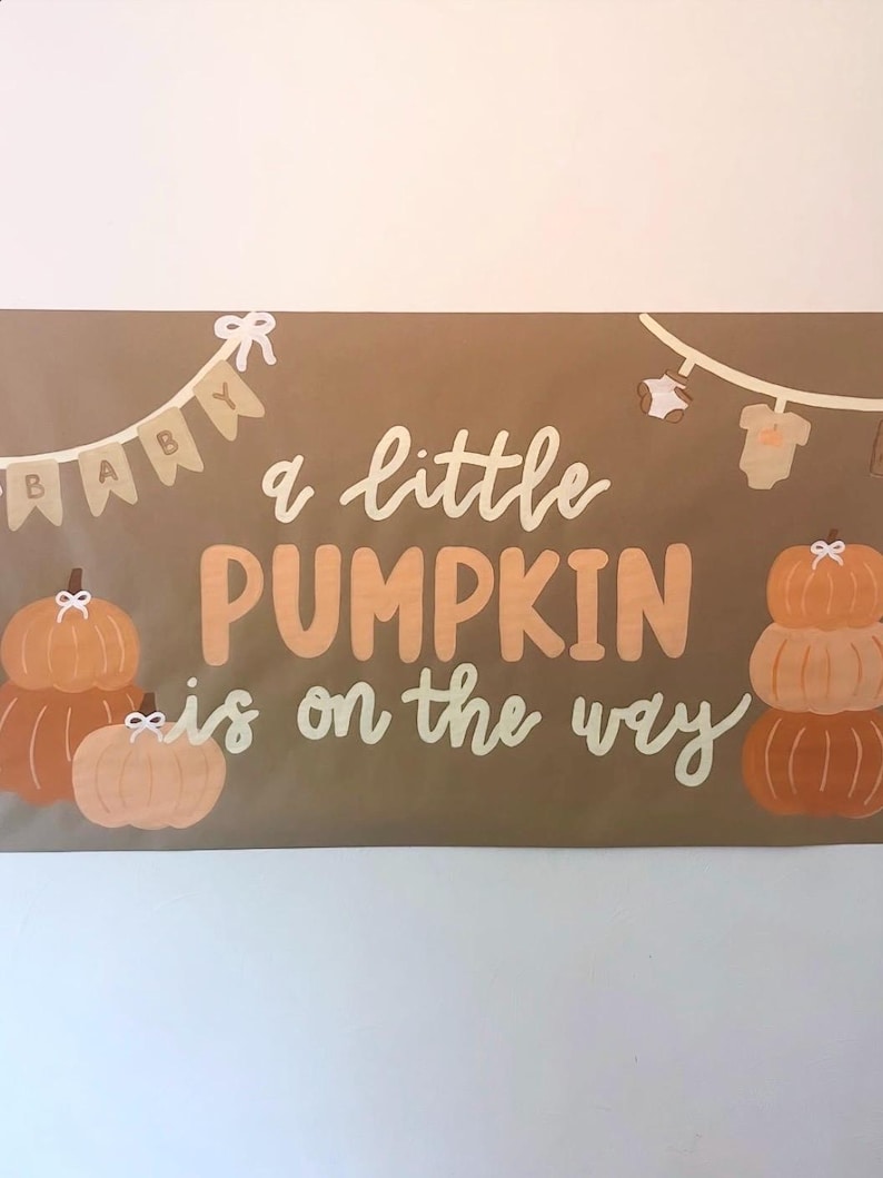 May include: A rectangular brown banner featuring the text "a little PUMPKIN is on the way" in white and orange. The design includes illustrations of orange pumpkins with white bows and a string of baby clothes and pennants with the word "BABY".