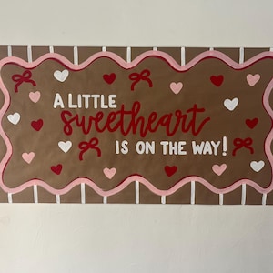 May include: A rectangular brown sign with a scalloped pink border and white vertical stripes. The text reads A LITTLE sweetheart IS ON THE WAY! in red and white. The design is decorated with small red, pink, and white hearts and red bows.