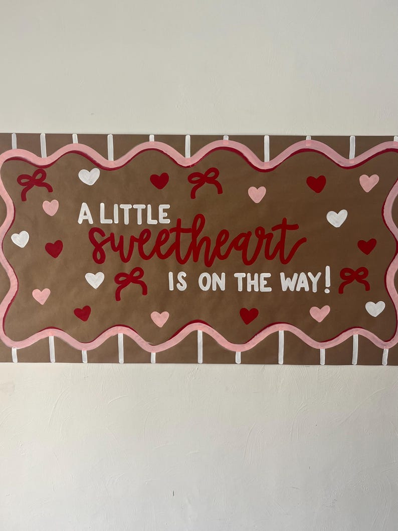 May include: A rectangular brown sign with a scalloped pink border and white vertical stripes. The text reads A LITTLE sweetheart IS ON THE WAY! in red and white. The design is decorated with small red, pink, and white hearts and red bows.