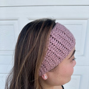 May include: A pink crocheted headband with a textured pattern. The headband is worn on a person's head.
