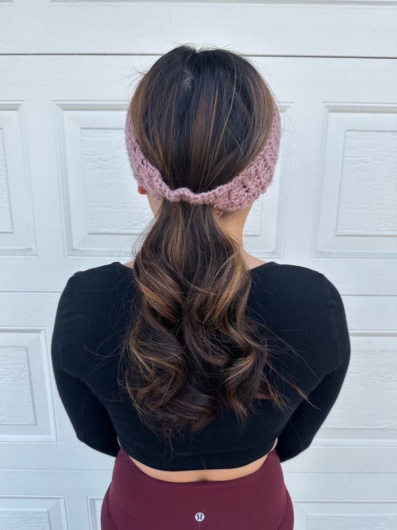 May include: A woman with long brown hair wearing a pink crocheted headband. Her hair is pulled back in a ponytail.
