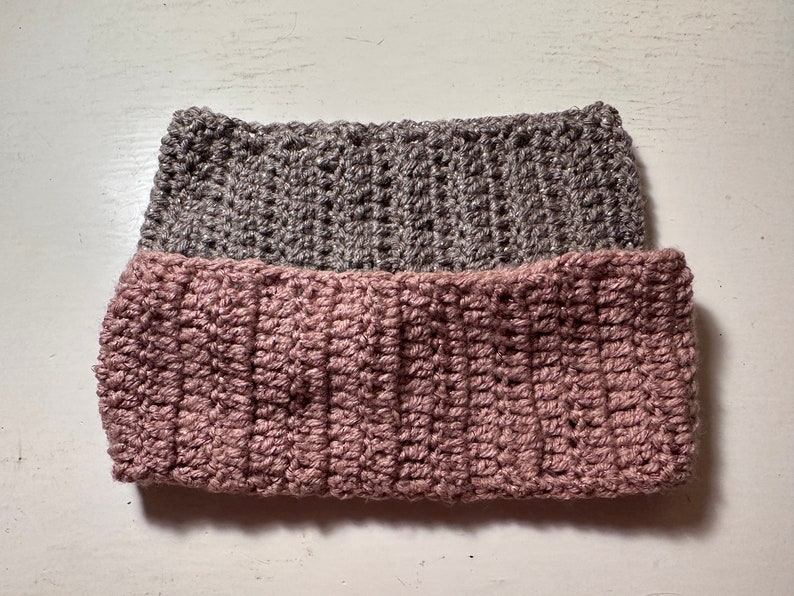 May include: Two crocheted headbands, one in a light pink color and the other in a gray color. The headbands are stacked on top of each other.